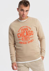 Felpa beige con texture a coste, scollo rotondo e stampa grafica arancione con la scritta "1986 CHICAGO VINTAGE SPORT". Maniche lunghe e vestibilità slim.