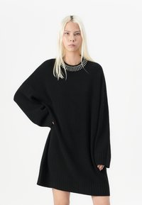 Svart strikket oversized kjole med ribbestrikket nederkant og mansjetter, med en naglet utringning. Teksturen ser myk ut, med en avslappet passform.