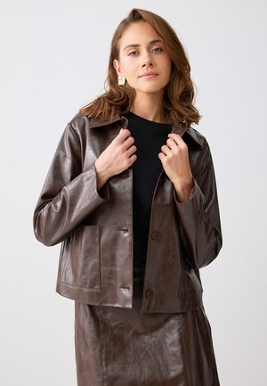 Femme aux cheveux bruns mi-longs, portant des boucles d'oreilles dorées, une veste en cuir brun foncé et une jupe assortie, ajustant le col de sa veste.