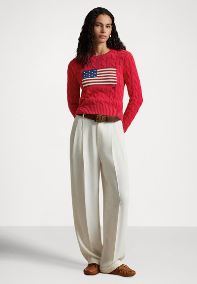 Pulover roșu tricotat cu model cu steagul american pe partea din față, asortat cu pantaloni albi cu croială largă și pantofi maro de tip mocasin.