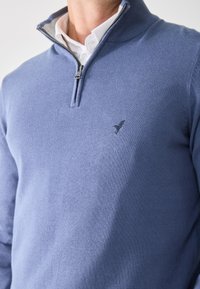Mann iført en lys blå quarter-zip genser over en hvit skjorte med krage, med en liten fuglelogo på brystet.