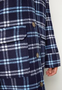 Tmavě modrý kostkovaný kabát s světle modrými a bílými kostkami, s klopovými kapsami a stříbrnými knoflíky, nošený na pravé straně osobou s viditelnou rukou.