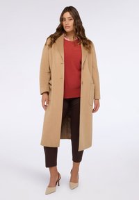 Cappotto lungo beige con design monopetto, ampi revers e due tasche frontali, indossato sopra un maglione rosso e pantaloni marroni.