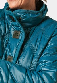 Gequiltete, tealfarbene Daunenjacke mit hohem Kragen, ausgestattet mit schwarzen Druckknöpfen und strukturiertem Nähen. Weiches, glänzendes Material.