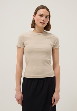 Vrouw met kort bruin haar, gekleed in een beige nauwsluitend T-shirt met korte mouwen en een zwarte rok, staand tegen een effen lichte achtergrond.