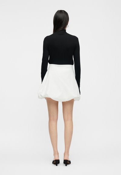Γυναίκα που στέκεται με την πλάτη γυρισμένη, φοράει μαύρο μακρυμάνικο τοπ, λευκή μίνι φούστα με όγκο και μαύρα mule με τακούνι, σε απλό λευκό φόντο.