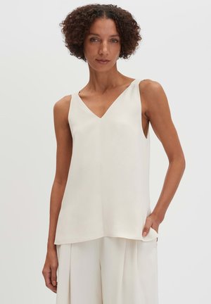 Femme aux cheveux bouclés portant un haut blanc sans manches à col en V et un pantalon large blanc assorti, debout avec une main dans la poche.