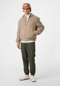 Beige bomberjakke med lynlås, ribbet krave og sidelommer, kombineret med olivengrønne joggers og hvide sneakers på en model.