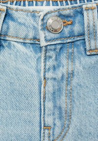 Světle modré džíny z denimu s kovovým knoflíkem, kontrastním oranžovým prošíváním a vyšisovaným vzorem v oblasti pásu a zipu.