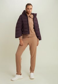 Giacca imbottita viola con cappuccio, top beige strutturato e pantaloni coordinati, abbinati a sneakers bianche, su uno sfondo chiaro.