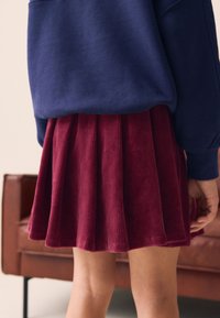 Gonna in velluto a coste color marrone scuro con pieghe, abbinata a una felpa blu scuro. Tessuto liscio; cuciture visibili lungo l'orlo.