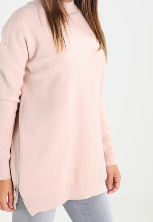 Femme portant un pull léger rose pâle à manches longues avec des fermetures éclair latérales, associé à un pantalon gris clair sur fond blanc.