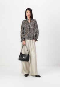 Δημιουργικό πουκάμισο με φουσκωτά μανίκια σε σκούρες αποχρώσεις, συνδυασμένο με ψηλόμεσα γυαλιστερά μπεζ παντελόνια και κρατώντας μια δομημένη μαύρη τσάντα.