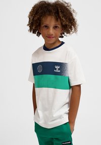 Göndör hajú gyermek, fehér, kék és zöld csíkos pólóban és zöld nadrágban, kezét zsebre dugva, előre nézve.