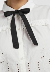 Camicia bianca con bottoni e cravatta nera al collo, con ricami decorativi e un dettaglio di rouches lungo il giogo.