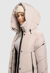 Giacca invernale imbottita di un beige chiaro con un grande cappuccio, zip nera e dettagli hardware, e tasche laterali. Tessuto texturizzato con design trapuntato.