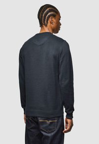 Hombre con el cabello trenzado de espaldas, vistiendo una camisa de manga larga oscura y jeans de mezclilla oscuros, contra un fondo claro y liso.