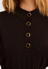 Czarna dzianinowa bluzka z wysokim kołnierzem ozdobionym trzema kwadratowymi, złotymi guzikami. Tkanina jest gładka o subtelnej fakturze, z elastycznym paskiem w talii.
