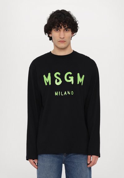 Black long-sleeve T-shirt with green brushstroke "MSGM MILANO" text. Made of cotton, featuring a crew neck and relaxed fit. Denim jeans worn below.