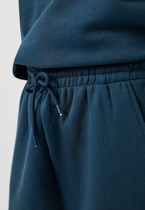 Pantaloni della tuta blu scuro con fascia elastica in vita e coulisse annodata, top in tessuto blu scuro parzialmente visibile.