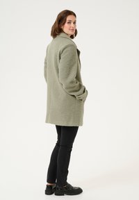 Lichtgroene, zacht aanvoelende, mid-length jas met een kraag, zijzakken en knoopdetails, gecombineerd met zwarte slim-fit jeans en schoenen.