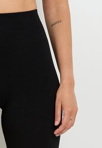 Leggings neri a coste con vita alta, caratterizzati da una texture liscia. Un tatuaggio è visibile sul braccio accanto a loro.