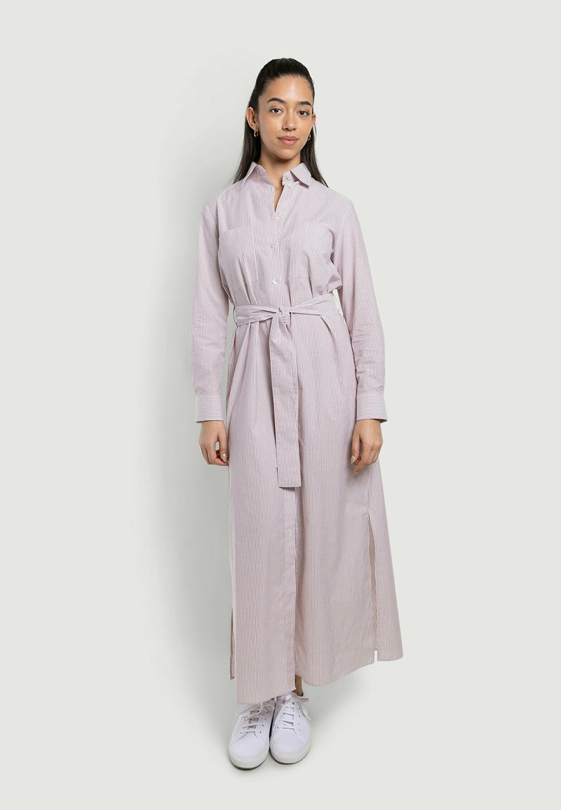 Femme aux longs cheveux foncés portant une robe longue à manches longues rayée rose clair ceinturée et des baskets blanches, debout sur un fond uni.