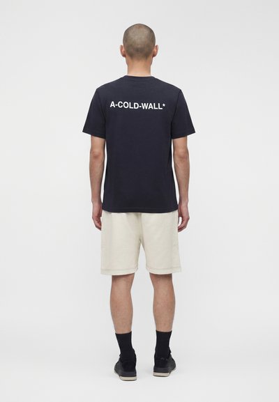 Άνδρας που στέκεται πλάτη, φοράει μαύρο T-shirt A-COLD-WALL*, κρεμ σορτς, μαύρες κάλτσες και μαύρα αθλητικά σε άσπρο φόντο.