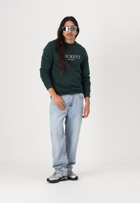 Zelená mikina s logom "Hackett London", svetlomodré džínsy a strieborné športové topánky. Model má na sebe reflexné slnečné okuliare.