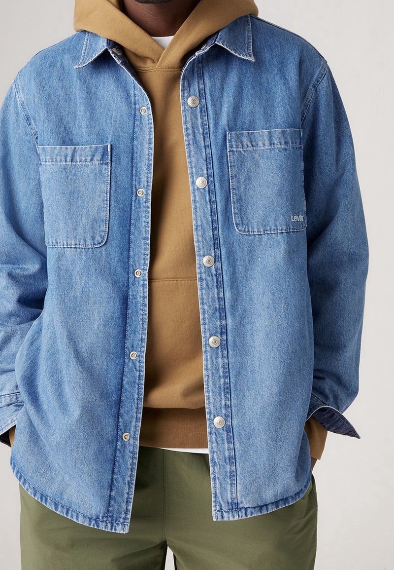 Jasnoniebieska koszula dżinsowa z dwoma kieszeniami na piersi i srebrnymi zatrzaskami, nałożona na brązową bluzę z kapturem, zestawiona z zielonymi spodniami.