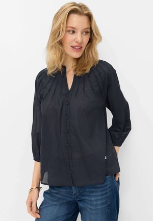 Donna con capelli biondi che indossa una camicetta nera abbottonata e jeans blu, guarda verso destra con un leggero sorriso, mano in tasca.