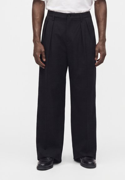 Mann trägt lockere schwarze Hose, schwarze Schuhe und ein weißes Hemd, steht vor einem schlichten hellen Hintergrund.