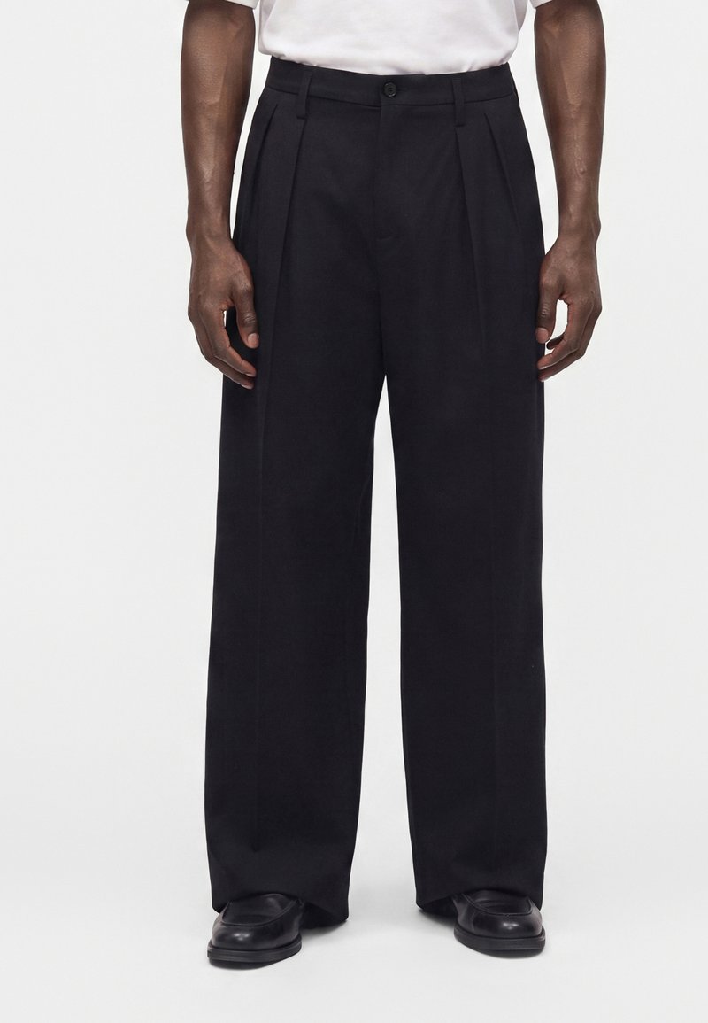 Hombre que lleva pantalones negros holgados, zapatos negros y una camisa blanca, de pie contra un fondo claro y liso.
