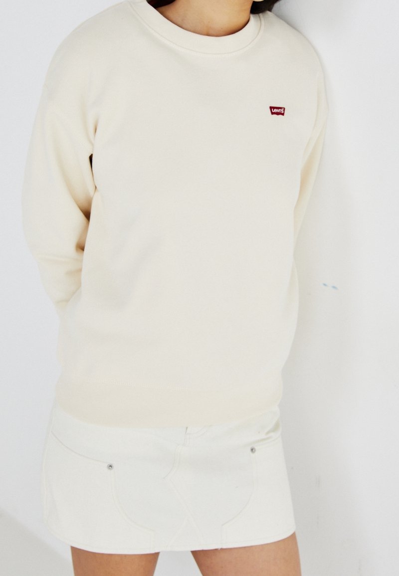 Personne portant un sweat crème avec un petit logo rouge Levi's et une mini-jupe crème assortie sur un fond blanc uni.