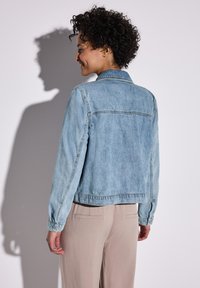 Chaqueta de mezclilla azul claro con mangas largas, puños abotonados y un cuello clásico. Presenta un yugo en la parte trasera y detalles cosidos a lo largo de las costuras.