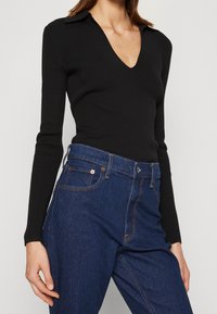 Black ribbed long-sleeve top with a deep V-neck, paired with dark blue high-waisted jeans featuring contrast stitching and classic pockets.