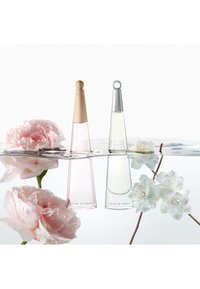 Dos frascos de perfume, uno rosa con una tapa de madera, el otro transparente con una tapa plateada, flotan en agua junto a flores rosas y blancas.