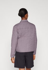 Prošívaná bunda světle purpurové barvy s límcem stojáčkem, žebrovanou strukturou a krátkým střihem. Kombinováno s černými šortkami, pohled zezadu.