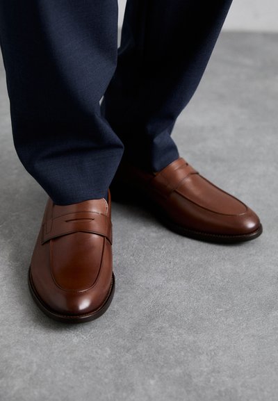 Mocasines de cuero marrón con un acabado suave, con detalles decorativos en la parte superior. Llevados con pantalones oscuros de color azul marino sobre una superficie gris.