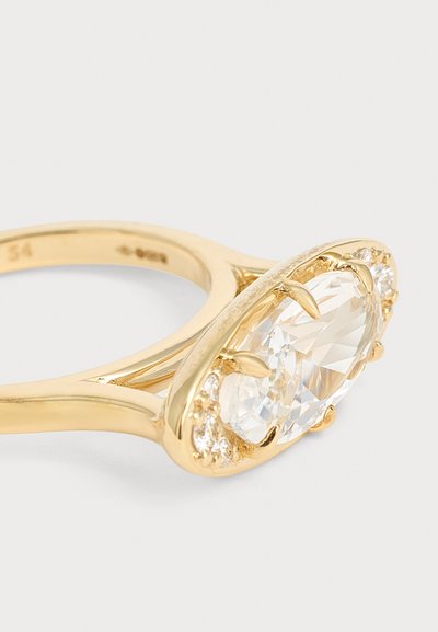 Bague en or avec une pierre précieuse claire ovale au centre, entourée de petites pierres claires rondes et maintenue par des griffes.
