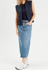 Frau in einer ärmellosen, gesteppten marineblauen Weste, schwarzem Top, hochgeschnittenem hellblauen Jeansrock, weißen Sneakers und einem silbernen Kettengürtel.