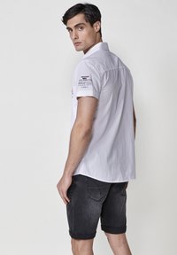 Camisa blanca de manga corta con cuello, que presenta texto negro en el hombro izquierdo. Combinada con pantalones cortos de mezclilla oscuros y remangados.