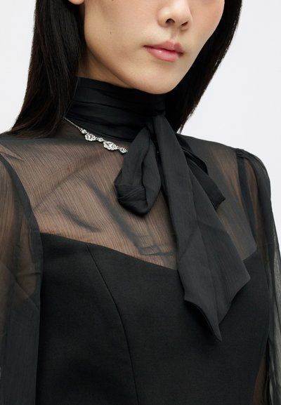 Γυναίκα φορώντας μια διαφανή μαύρη μπλούζα με φιόγκο στον λαιμό και ασημένιο κολιέ με διάφανες πολύτιμες πέτρες.