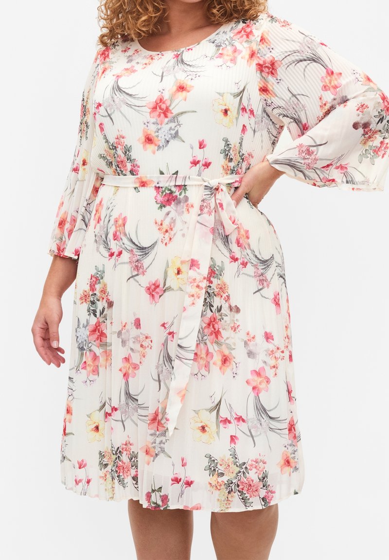 Robe fleurie longueur genou en tissu léger plissé, fond crème, ornée de fleurs roses et jaunes, avec une ceinture en tissu.