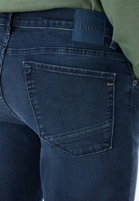 Jeans en denim bleu foncé avec une surface texturée, dotés d'une patch sur la ceinture avec le logo "Brax", et d'une poche arrière avec une couture subtile.