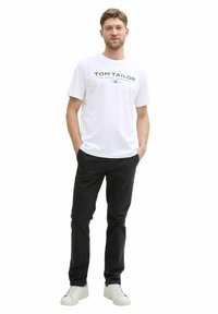 Weißes Baumwoll-T-Shirt mit schwarzem Text-Logo, kombiniert mit schwarzen Hosen und weißen Turnschuhen. Einfaches Design, lässige Passform und keine sichtbaren Muster.