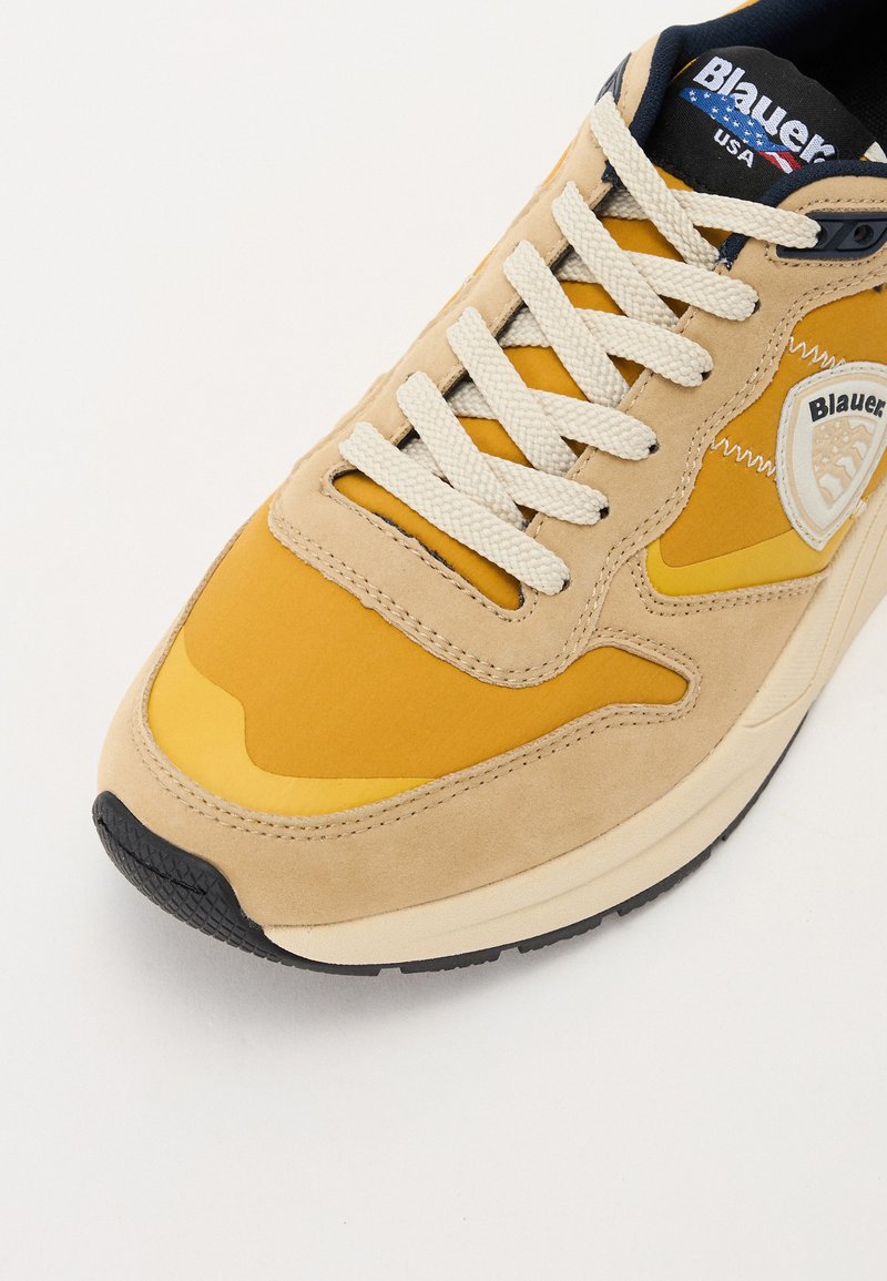 Beige und gelbe Sneaker mit einer strukturierten Wildleder-Oberfläche, cremefarbigen Schnürsenkeln und einer schwarzen Gummisohle. Logodetail an der Seite.