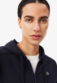 Navy zip-up hoodie with textured fabric, featuring a white inner layer, kangaroo pocket, and green logo accent on the chest.