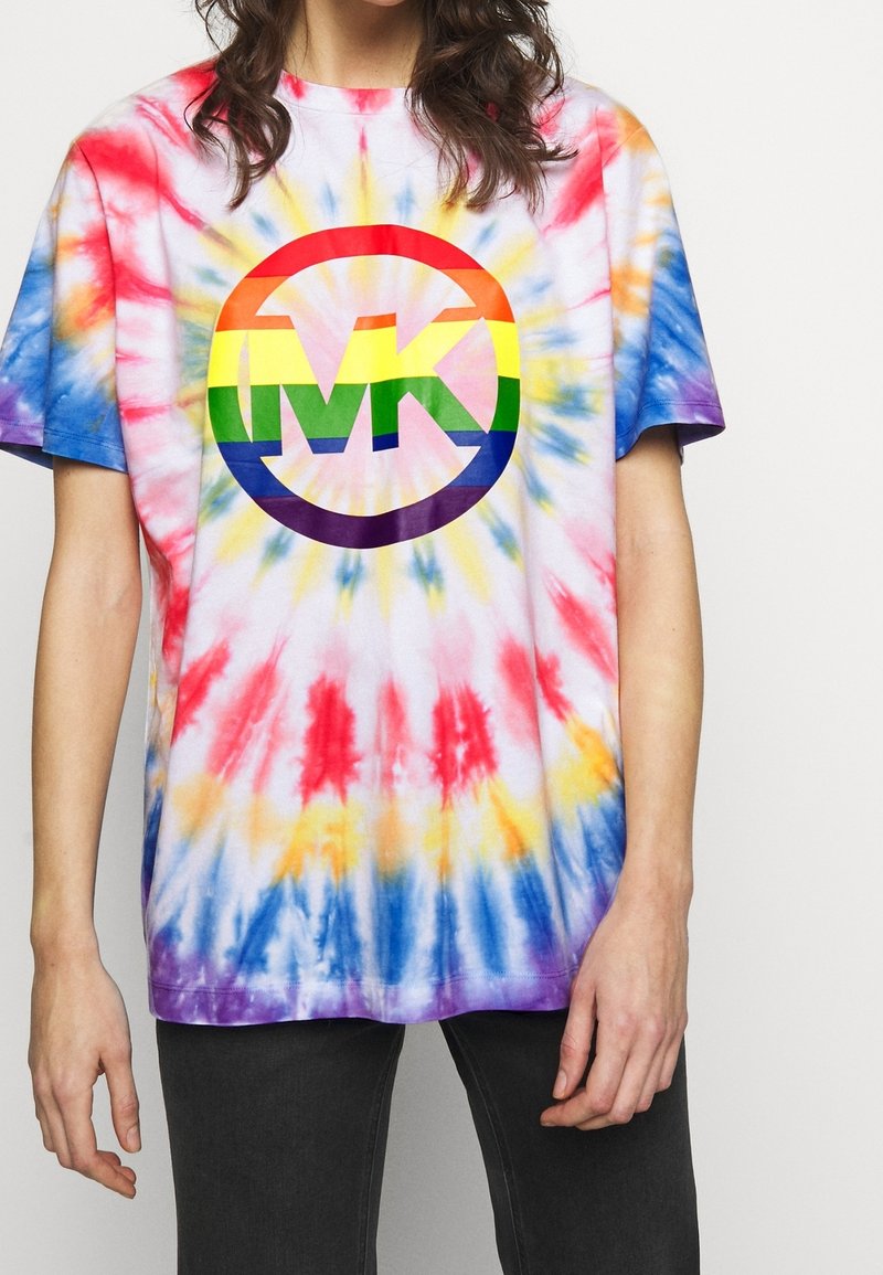 Personne portant un T-shirt tie-dye arc-en-ciel avec un grand logo MK aux couleurs de l'arc-en-ciel sur la poitrine, associé à un pantalon foncé.