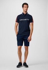 Marineblauwe polo met witte en oranje accenten, voorzien van een logo op de voorkant, gecombineerd met marineblauwe shorts en zwarte sneakers.